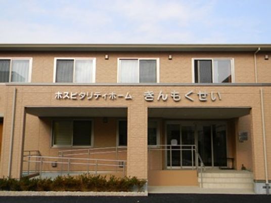 ブラウン系の建物は２階建て。周りには緑が植えられ、外壁にはホーム名が書かれている。