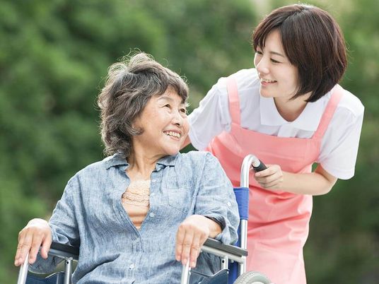 デニムシャツを着た高齢女性が車いすに座り、エプロンを付けた女性スタッフが付き添い、散歩をしている。