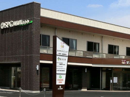 ブラウン系の外壁の２階建ての建物の玄関には自動ドアが設置され、２階にはベランダが付き、館銘板もある。