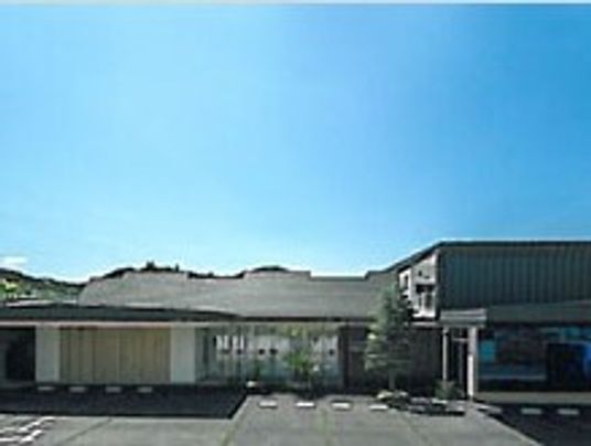 青い空の下に建っている平屋建ての建物。広い駐車場があり、周辺には緑もあふれている。
