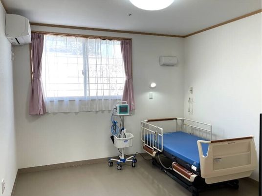 ベッドサイドには緊急通報装置があり、介護ベッドやエアコン、医療器具も置かれている部屋。