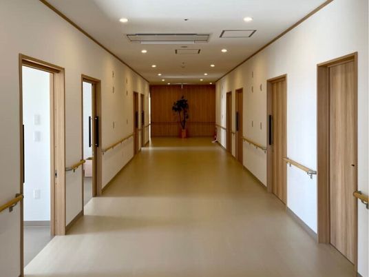 まっすぐな廊下は幅が広く、両サイドに手すりが付き、居室のドアが開いている。奥には観葉植物が飾られている。