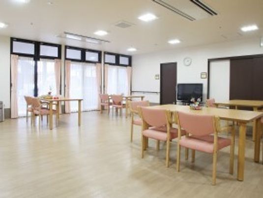 食堂にはピンクの椅子がセットされた木製のダイニングテーブルセットが複数置かれ、大きなテレビもあり、カーテンが付いた窓がたくさんある。