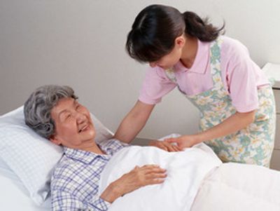白髪の高齢の女性入居者がベッドに横たわり、エプロンを付けた女性スタッフと笑顔で話している。