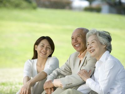 高齢夫婦と若い女性が日当たりのいい芝生広場で並んで座り、楽しそうに話をしている。