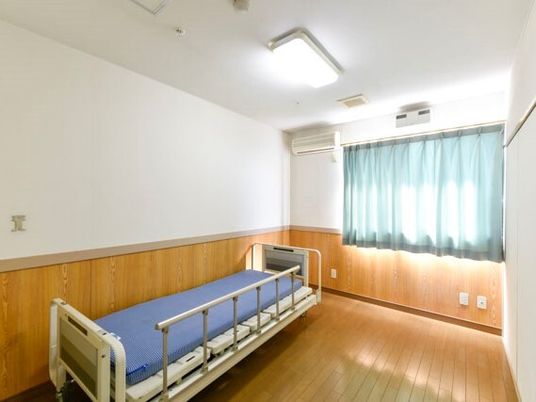 介護ベッドが壁際に置かれたフローリングの居室にはエアコンや照明が付き、窓にはカーテンが付いている。日当たりのいい部屋。