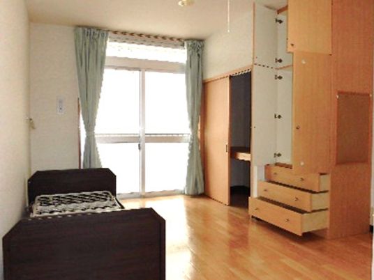 寝具付きのベッドが置かれたフローリングの部屋。収納スペースが多くあり、カーテン付きの窓も付いている。