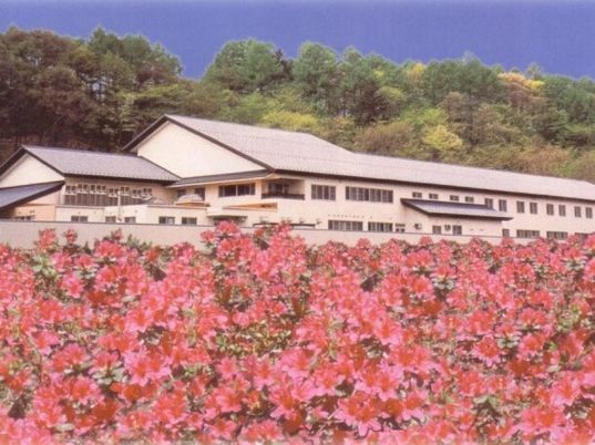 ピンクのつつじの花がたくさん咲いていて、その奥には白い屋根付きの建物があり、その奥には山がある。