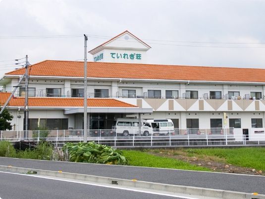 白い2階建ての建物にはオレンジの屋根や広い駐車場が付き、周辺には畑が広がっている。