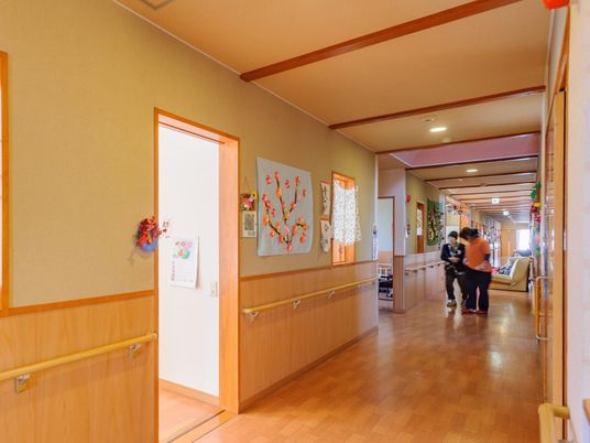 まっすぐな廊下を歩いている高齢女性にスタッフが付き添っている。廊下には壁画が飾られ、手すりも付いている。
