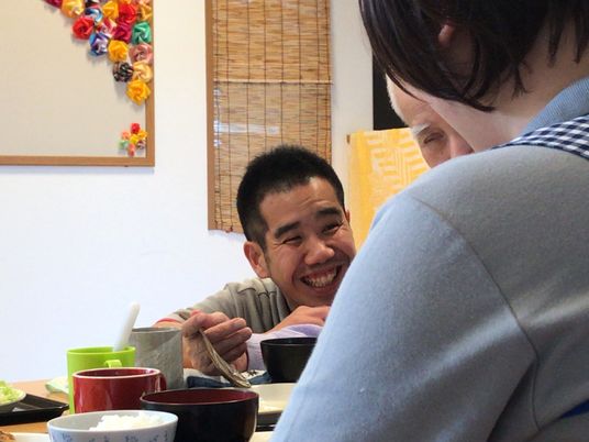 男性スタッフが高齢男性のそばで笑顔で話を聞いている。エプロンを付けた女性スタッフもそばにいる。