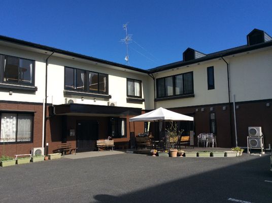 1階部分はブラウン、2階部分は白い2階建ての建物はエル字型で、パラソル付きテーブルセットも敷地内にある。