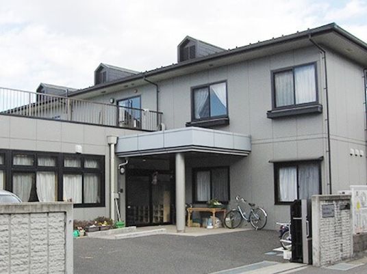 グレーの２階建ての建物。玄関周りには自転車などが置かれている。