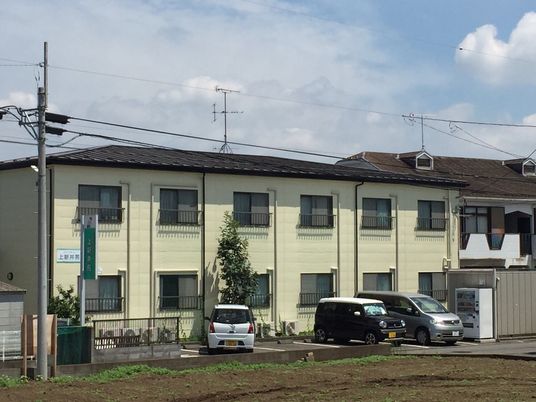 ２階建ての建物は日当たり良好でベージュ系の外壁。敷地内には駐車場があり、車が止まっている。