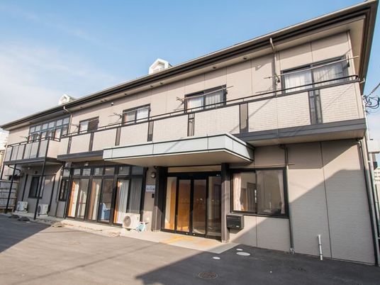 日当たりのいい場所に建っているベージュの外壁の2階建ての建物。玄関に屋根もある。