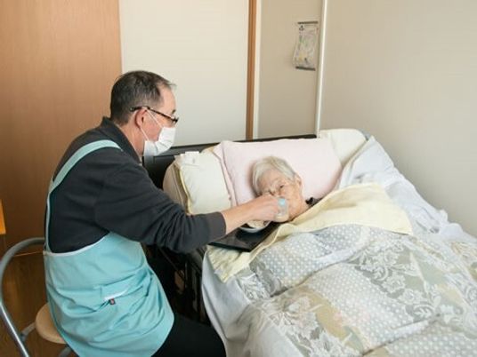 ベッドに横になる高齢女性の水分摂取を介助している男性スタッフ。