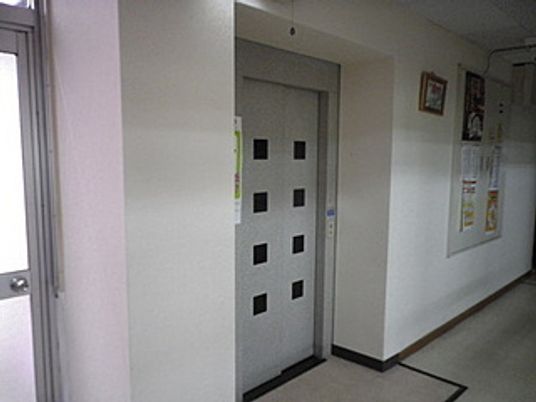 グレーの扉のエレベーターが設置されている館内。左側にはドアがあり、右側には掲示物がある。