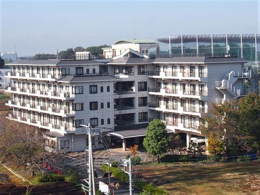 白をメインとしたエル字型の建物は５階建て。周辺には緑が多く、駐車場もある。