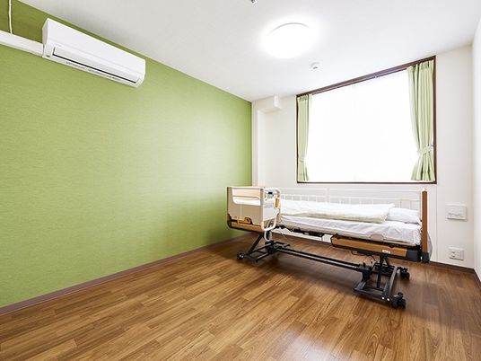 グリーンの壁紙にフローリング、介護用ベッドとエアコン、照明がある個室