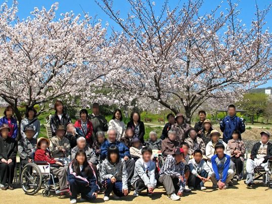 桜の木をバックに入居者とスタッフ、子供たちで撮影。青空が広がっている。