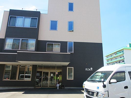 ベージュとダークグレーの建物にはたくさんの窓があり、駐車場には車が止められ、玄関には車寄せが付いている。