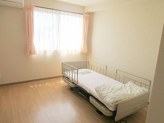 寝具付きのベッドが置かれたフローリングの部屋にはレースカーテンとカーテンが付いた大きな窓がある。