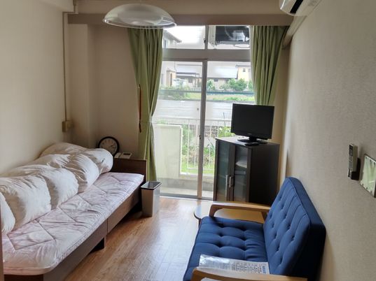 ブルーのソファや寝具つのベッド、テレビやエアコンなどがあるフローリングの部屋には大きな窓が付いている。