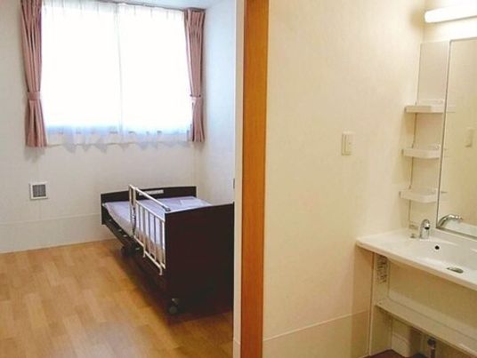介護ベッドが置かれたフローリングの部屋には洗面所があり、レースカーテンとカーテンが付いた大きな窓がある。