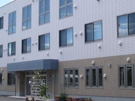 3階建ての建物。グレー系の色でまとめられ、玄関には屋根があり、低木も植えられている。