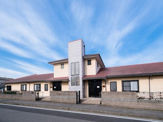 青い空の下に建っている2階建ての建物。エンジ色の屋根が付き、玄関の前にはブロックが敷かれ、敷地は塀で囲まれている。