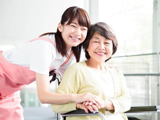黄色いカーディガンを着た車いすの高齢女性のそばにはピンクのエプロンを付けた若い女性スタッフがいて、二人とも笑顔。