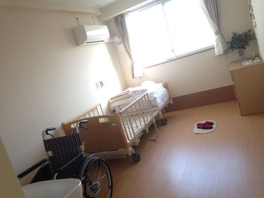 フローリングの居室。介護ベッドやエアコン、タンス、洗面所、車いすが置かれ、日当たり良好。