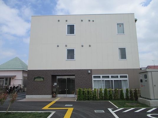 白とブラウンの３階建ての建物の敷地内には駐車場があり、点字ブロックも敷かれている。