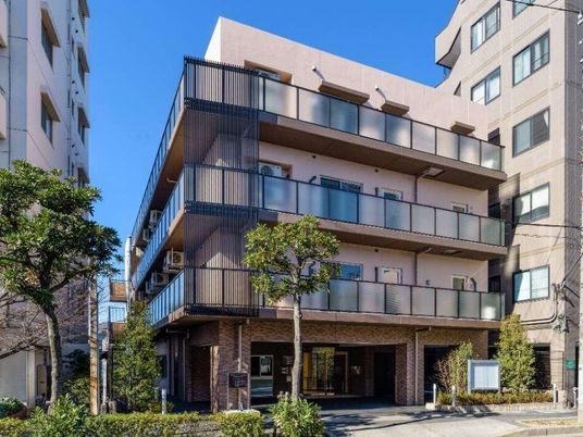 4階建ての建物はベージュで各フロアにベランダがあり、周辺には木も植えられている。