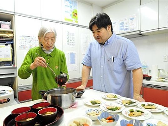 男性スタッフに見守られ、キッチンで汁物を取り分けている女性入居者。