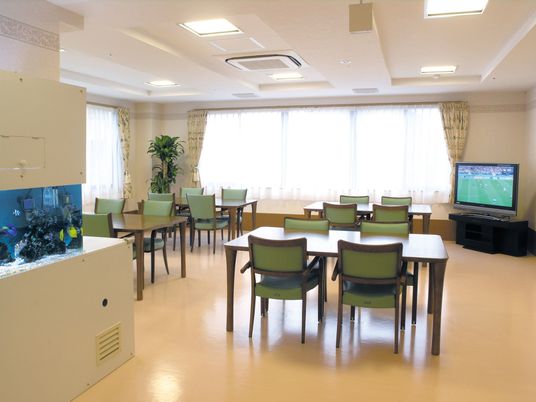 グリーンのいすが並べられた４人掛けのテーブルが複数並べられ、テレビスペースもある明るい食堂。