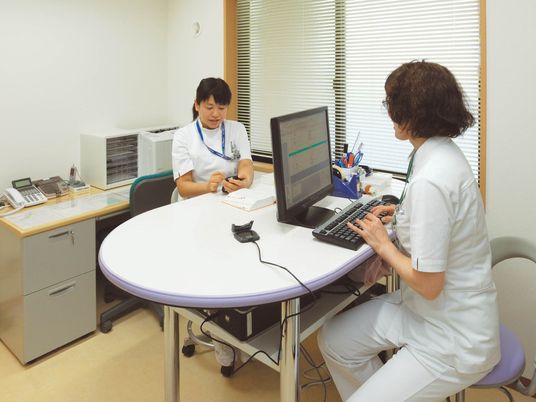 白衣を着た看護スタッフが2名話し合いをしている様子。一人はメモを取り、一人はパソコンを見ている。