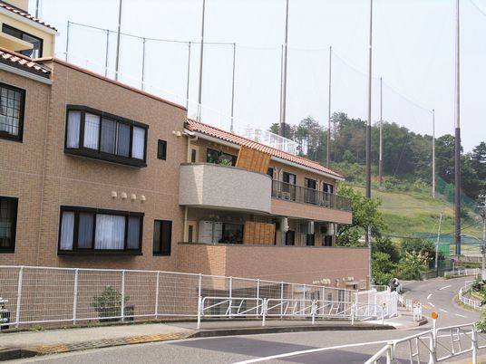 自然に囲まれたのどかな風景。奥には山や畑があり、手前に白いフェンスに囲まれたホームがある。