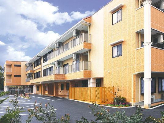 オレンジ色の建物の前には広い駐車場があり、玄関周辺には多くの木が植えられている。