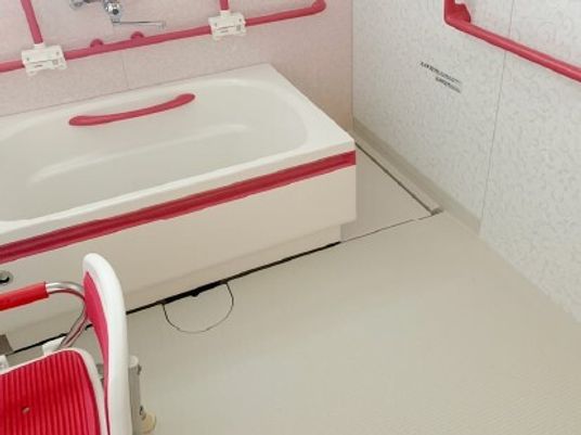真っ赤な手すりやシャワーチェアが設置された白を基調とした浴室。洗い場が広く、浴槽は可動式。