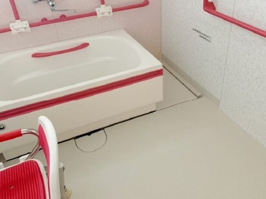 真っ赤な手すりやシャワーチェアが設置された白を基調とした浴室。洗い場が広く、浴槽は可動式。