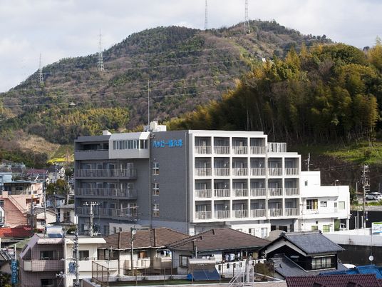 民家に囲まれたグレー系の大きな建物の外壁にはホーム名が書かれている。周りには山もある。