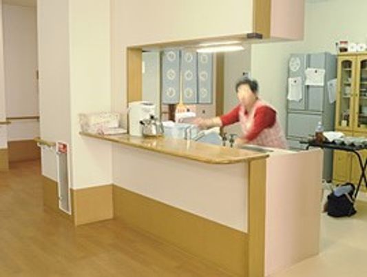 対面式キッチンでエプロン姿のスタッフが作業している姿が見える。キッチンには冷蔵庫や食器棚がある。