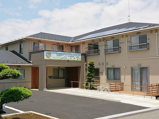 グレーの屋根が付いた２階建ての建物はベージュ系の外壁でエル字型。駐車場も設置されている。