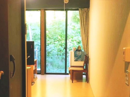 緑あふれる風景が大きな窓の外に広がっている。カーテンは開けられ明るい雰囲気の居室内には、いすなどの私物が持ち込まれている。