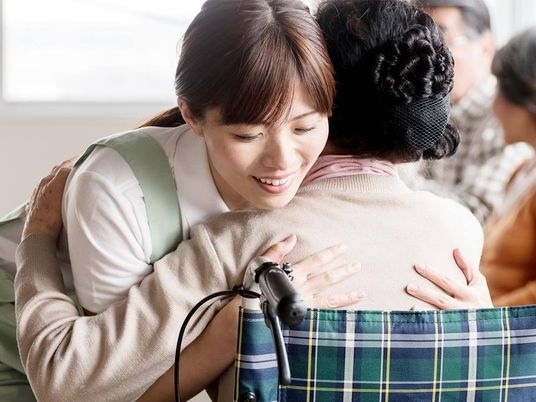 長い髪を後ろで一つに結んだ女性スタッフが、チェックの車いすに座った高齢女性を抱きかかえるようにして介助している様子。