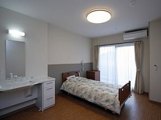 寝具付きのベッドやレースカーテンとカーテンが付いた大きな窓がある部屋にはワイドタイプの車いす対応の洗面所がある。