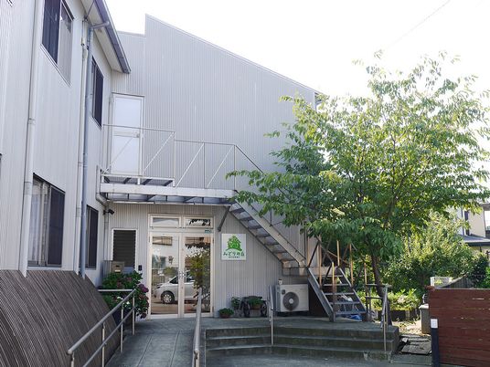 ２階建ての建物は穏やかな雰囲気。階段の横には木が植えられ、なだらかなスロープが設置されている。