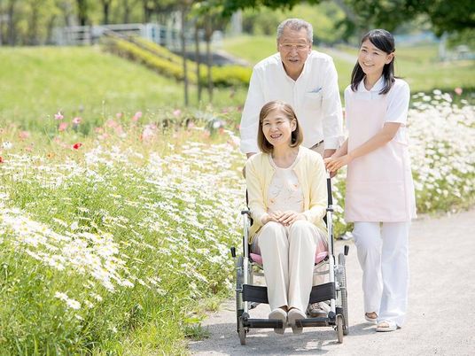 白い花がたくさん咲いている道を楽しそうに散歩している車いすの高齢女性。高齢男性とエプロンを付けた女性スタッフが付き添っている。