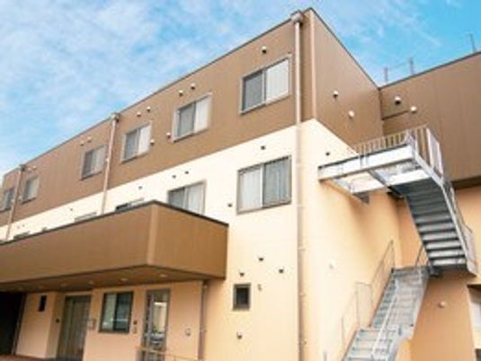 ブラウンとベージュの２階建ての建物は玄関に屋根があり、屋外階段が設置されている。